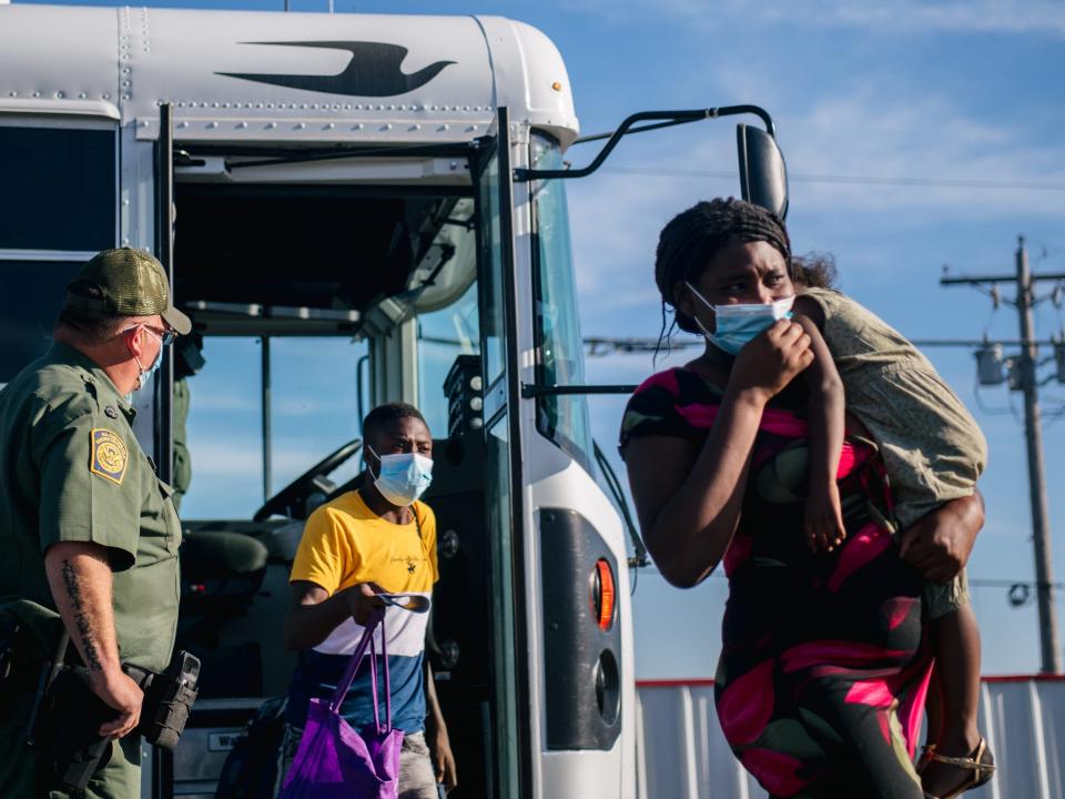 A woman wearing a face mask and carrying a child exits a bus, a man behind her in a face mask also exits the bus carrying a purple bag. A uniformed border patrol officer stands by the bus door watching them.