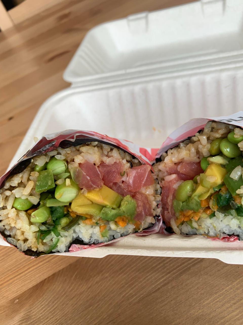 A build-your-own sushi burrito is made with rice, spicy tuna, carrot, edamame, avocado and seaweed, all wrapped in seaweed. Toppings are tamari, signature poke sauce and tempura flakes.