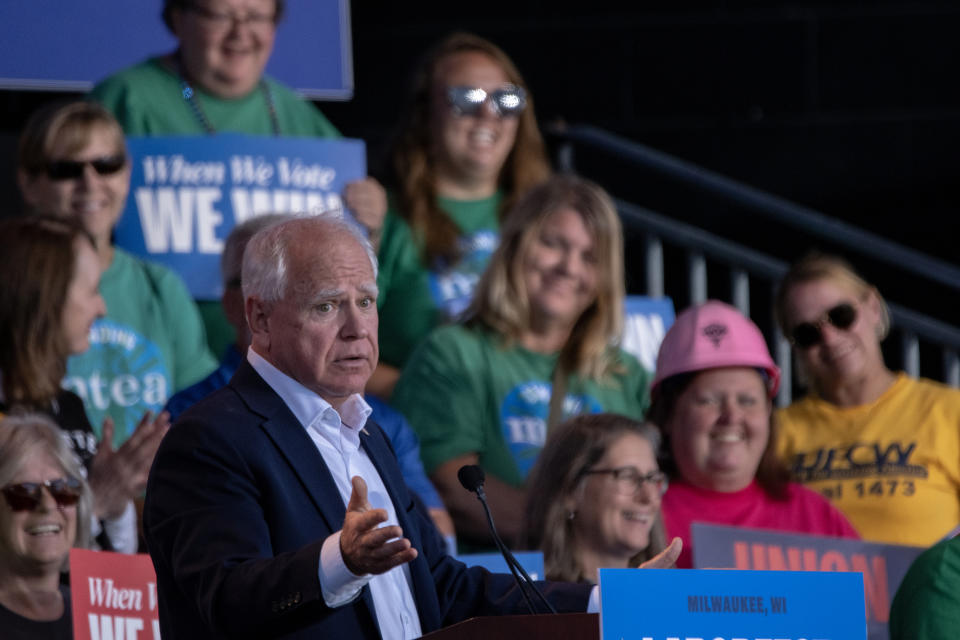 Tim Walz speaks at an outdoor rally surrounded by supporters holding signs. Some attendees wear shirts promoting voting and union support