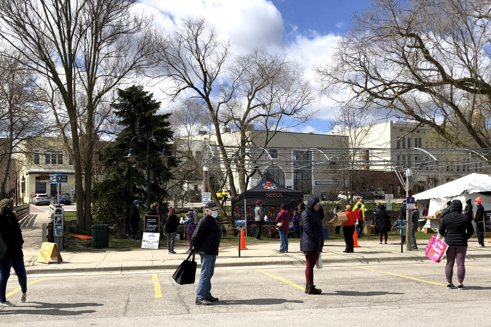 In this on May 9, 2020 photo, customers wait in a parking lot to pick up vegetables, baked goods and other items from the downtown farmers' market in Traverse City, Mich. In normal times, the lot would be crowded with shoppers browsing leisurely among dozens of stalls. But because of the coronavirus pandemic, they are asked to wear masks and observe social distancing while lining up for pre-ordered bags of food. (AP Photo/John Flesher)