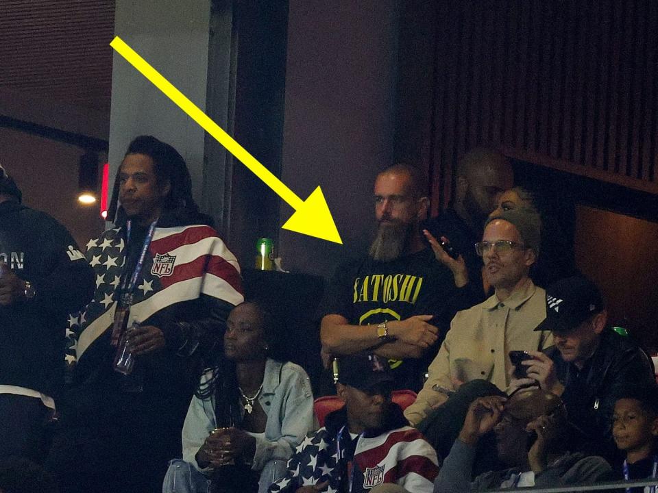 Group of people watching a sports game from the stands, with a skitched yellow arrow pointing out a t-shirt