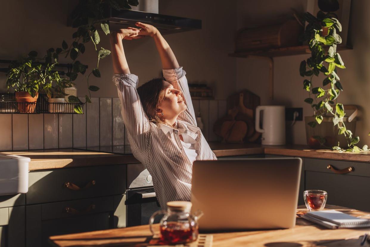 happy relaxed young woman sitting in her kitchen with a laptop in front of her stretching her arms above her head and looking out of the window with a smile