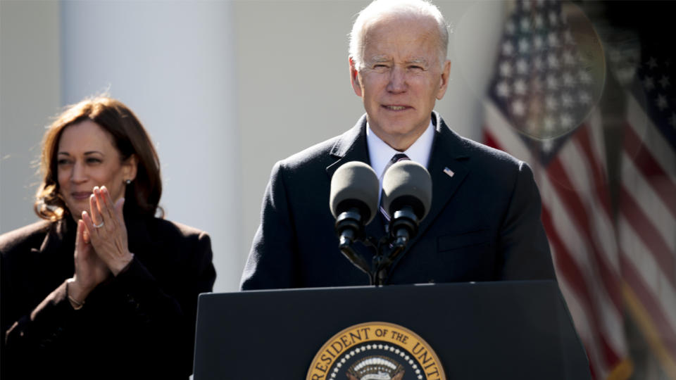 President Biden grimaces at the podium, as Vice President Kamala Harris applauds, behind him to his right.