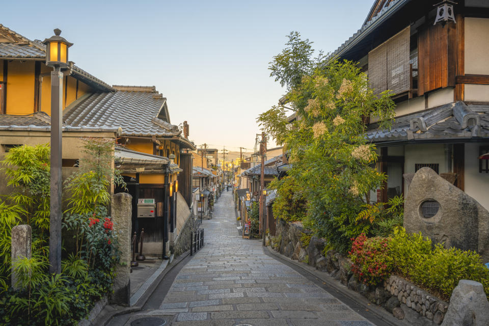 A narrow, stone-paved street in a quiet Japanese town, lined with traditional wooden buildings and lush greenery on either side