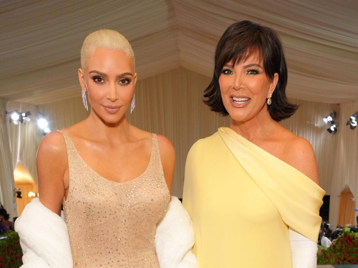 kim kardashian and kris jenner standing in a tent lined with flowing beige fabric on the sides and a red and blue carpet. kim is wearing a light beige dress with rhinestones all over and a white fur shawl, while kris is wearing a yellow dress that's open on one shoulder and white gloves. both are smiling