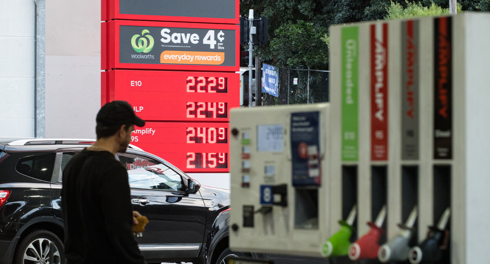 A driver returning to his car at a gas station, with a bowser in the foreground and a sign in the background showing fuel prices.