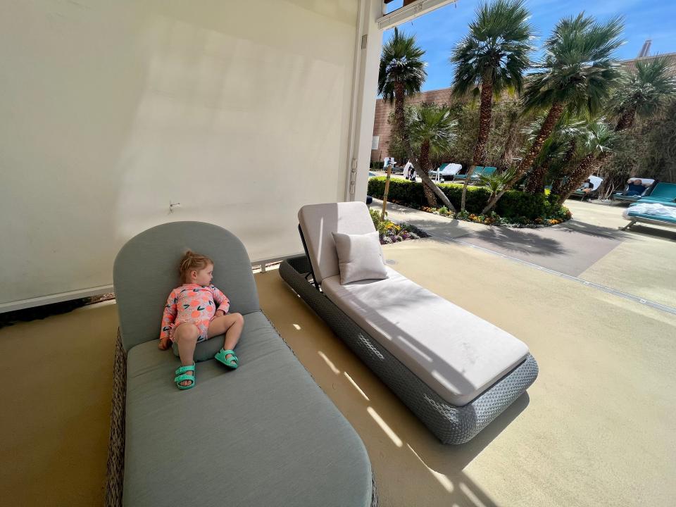 A child sits on a plush daybed next to an empty one in a pool area.