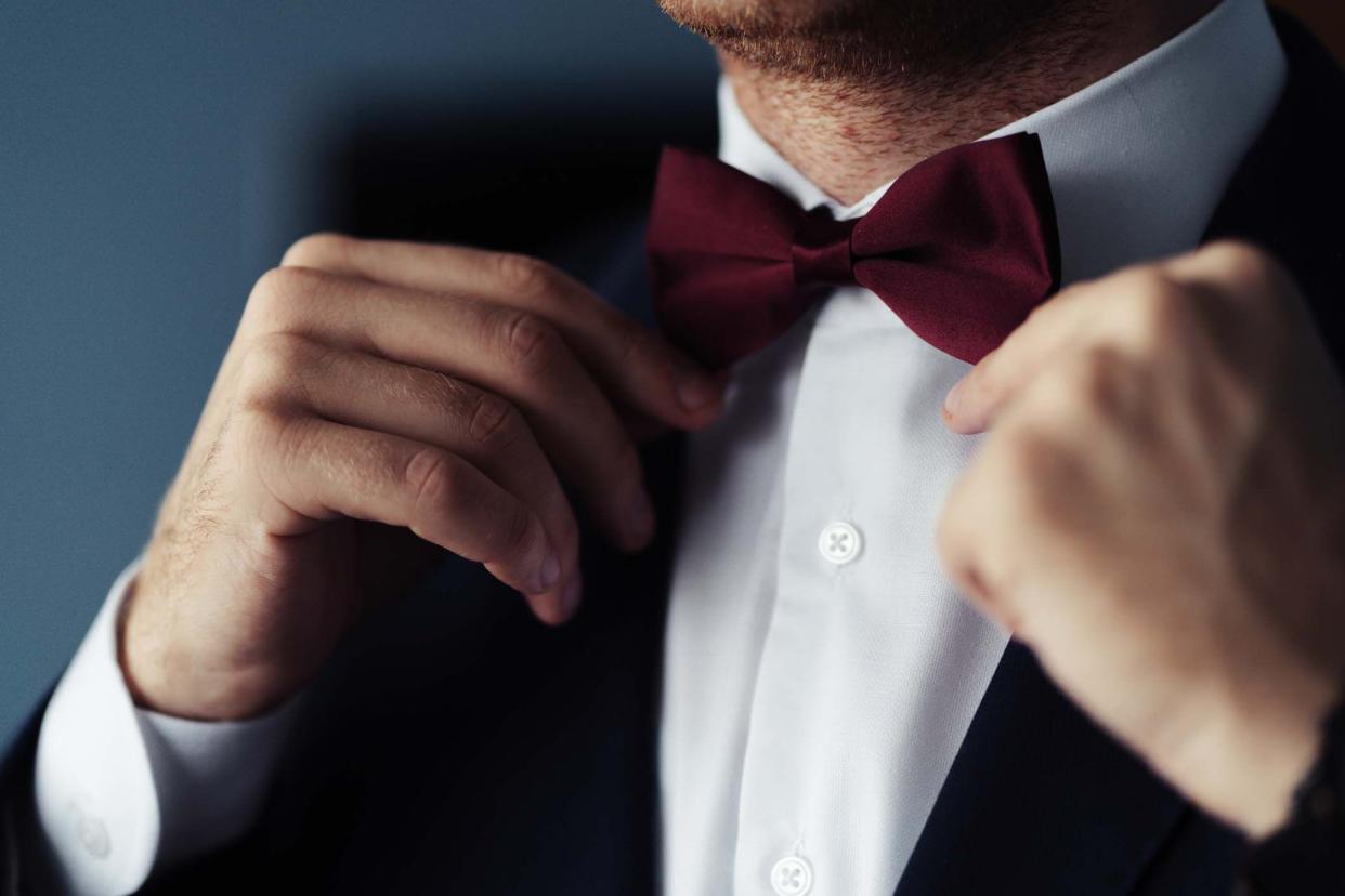 Closeup of man's hands fixing top front of tuxedo while wearing it, maroon bow tie and white button up shirt, with a blurred grey background