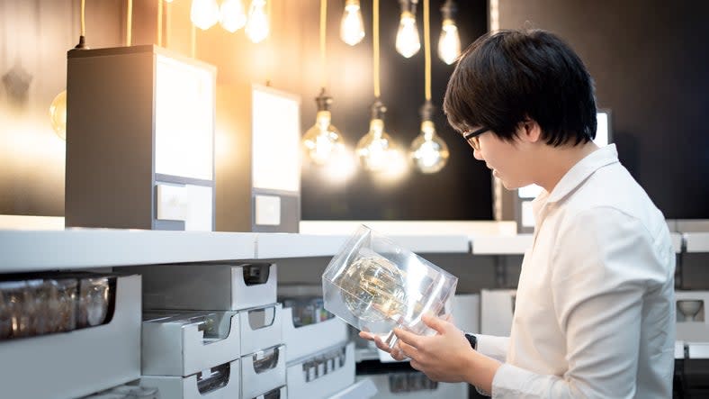 young-person-in-white-shirt-looks-at-a-light-fixture-on-an-aisle-in-a-retail-store-with-other-light-fixtures-on-display-and-in-boxes-on-shelves