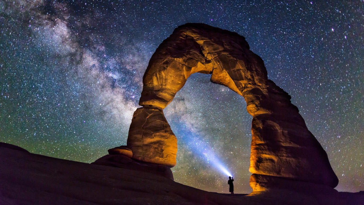  A person is standing in the middle of the Delicate Arch, Utah, USA, watching the milky way and lightning up the milky way with a flashlight beam. 