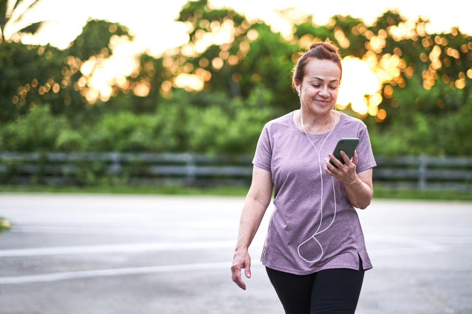 A woman in casual exercise attire, with earphones, walks outdoors while looking at her phone. Trees and a fence are blurred in the background