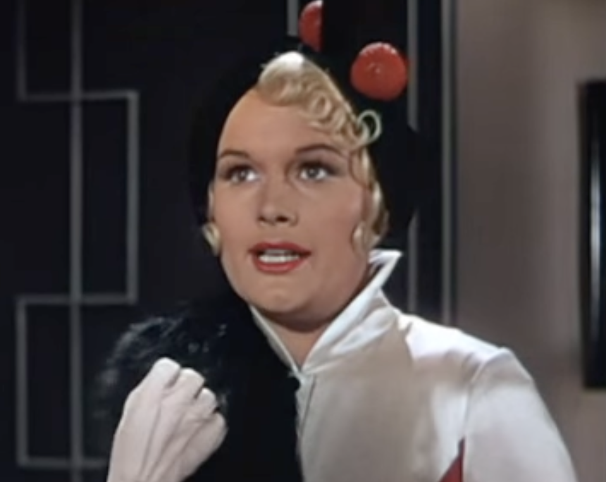 A woman in a vintage film wearing a high-necked outfit and a hat decorated with spherical ornaments holds a fur stole