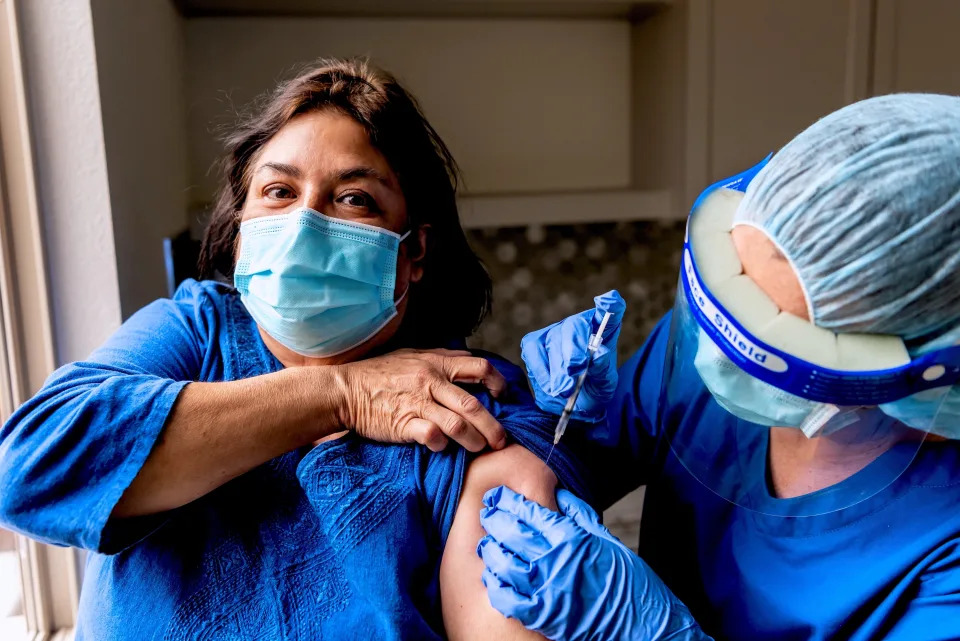 A woman with a blue mask on is given a COVID-19 shot by a healthcare worker. Both people are wearing blue shirts, and the healthcare worker wears a mask and face shield.