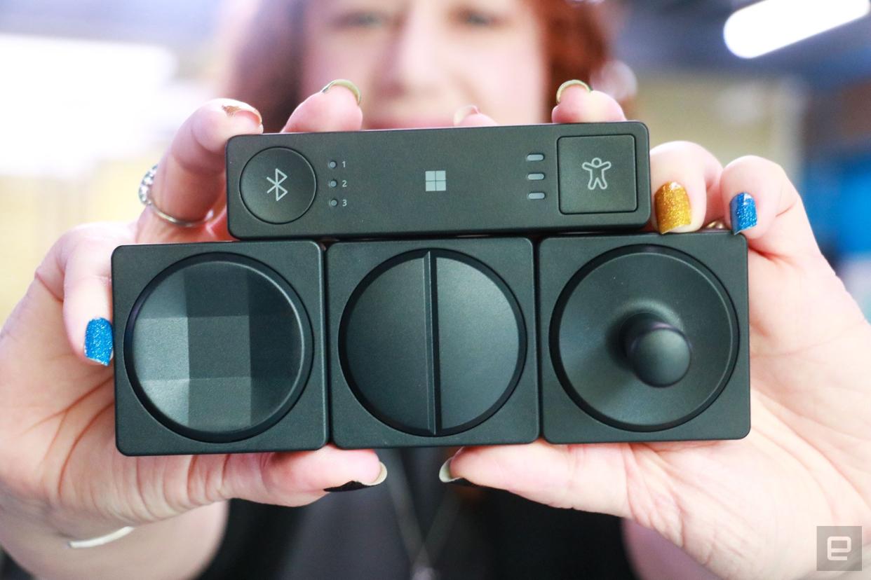 Three Microsoft Adaptive Buttons and an Adaptive Hub on top of them, held in mid-air by two hands with colorful nails. The Buttons have different toppers. From left to right, they are the eight-directional d-pad, the dual button and a joystick.