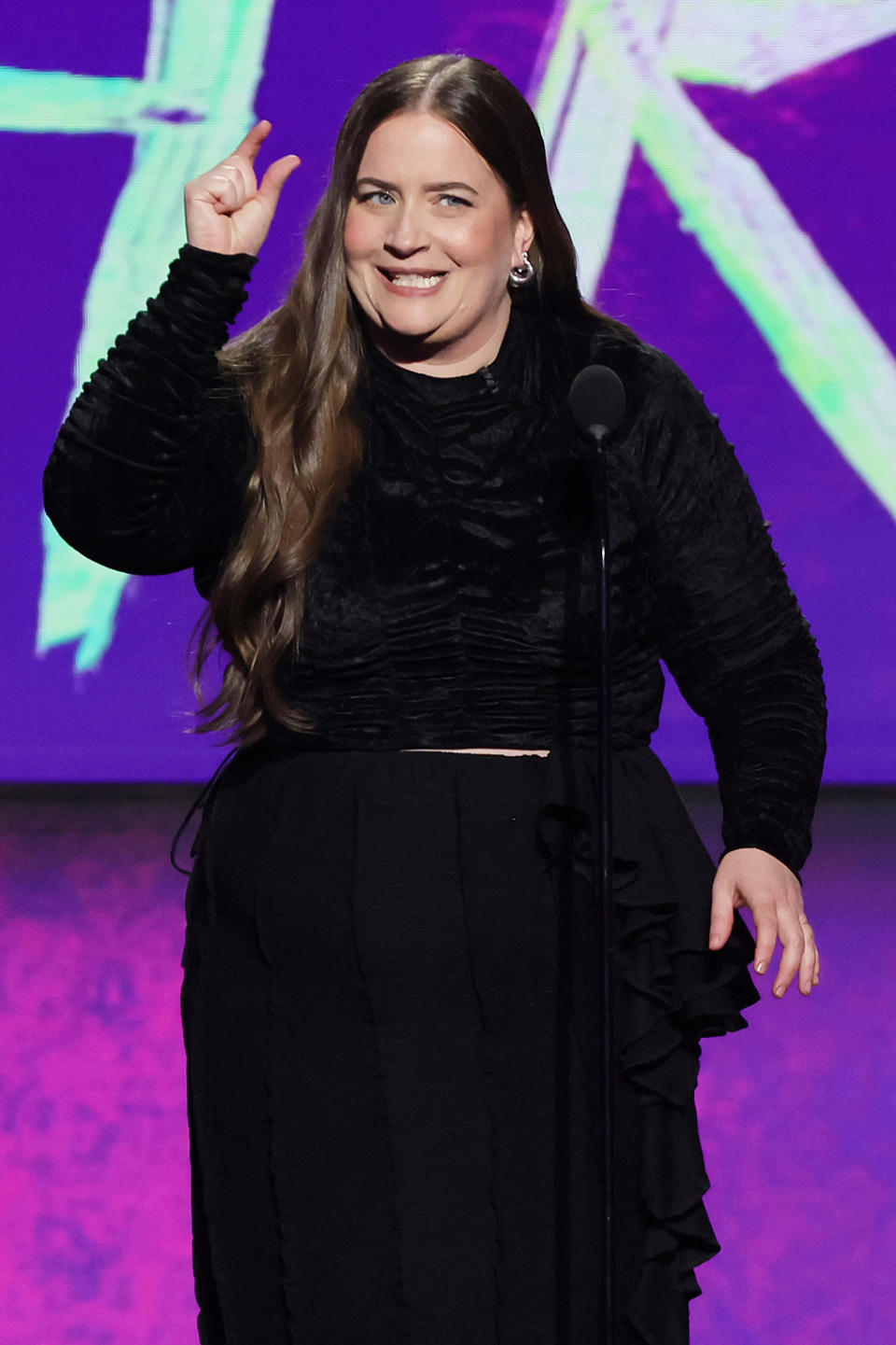 Aidy on stage wearing a long-sleeved top and skirt, smiling at the audience