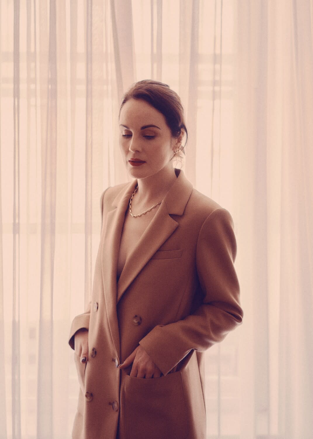A woman stands in front of sheer curtains with her hands in her coat pockets.