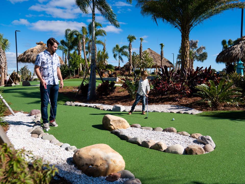 An adult and child playing mini golf on a bright blue day.