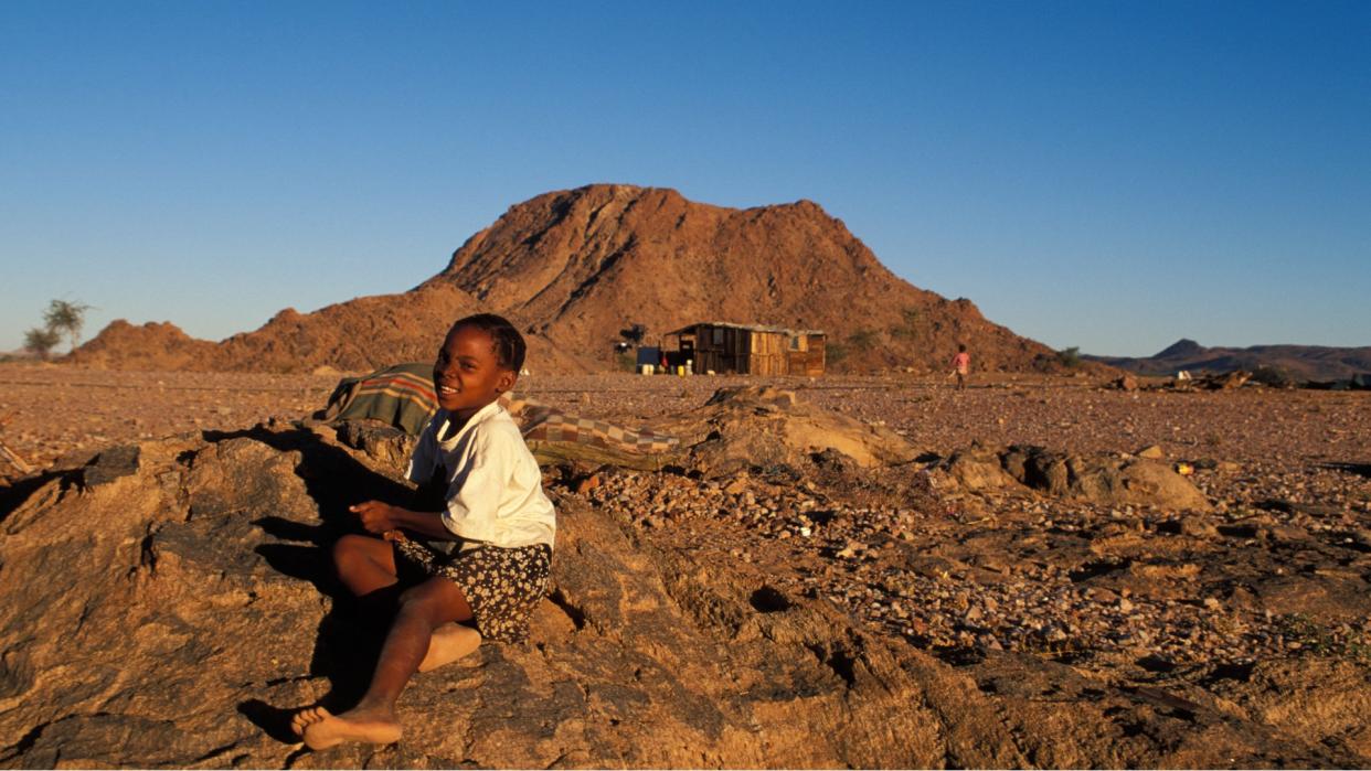  A Nama girl in Northern Cape province, South Africa. She wears a white shirt and a black skirt with white spots. She sits on the rocky ground in front of a small mountain in the background. 