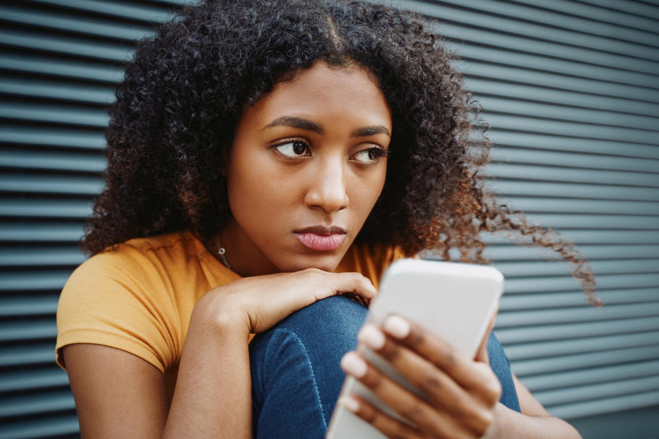Woman with curly hair and a pensive expression holding a smartphone while sitting against a metal shutter