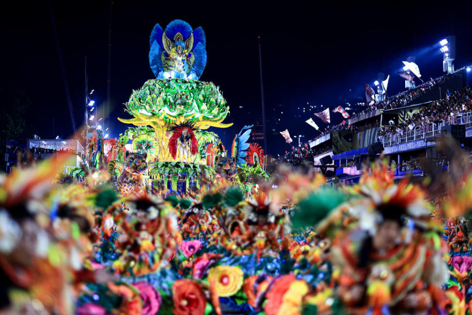 A vibrant, colorful float adorned with intricate decorations and peacock feathers partakes in a nighttime parade at the Rio Carnival, surrounded by dancing performers