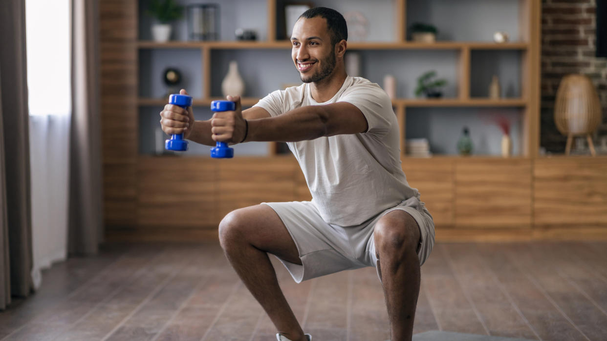  Man squats down at home holding dumbbells out in front of him. 