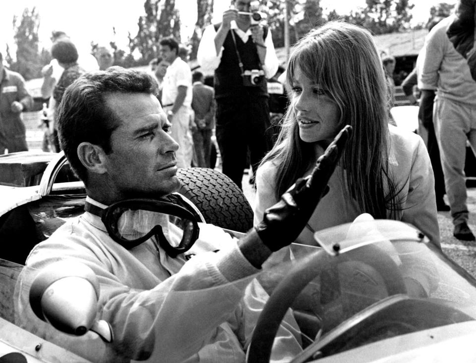 James Garner sits in a race car wearing driving gloves and goggles around his neck as Françoise Hardy appears alongside