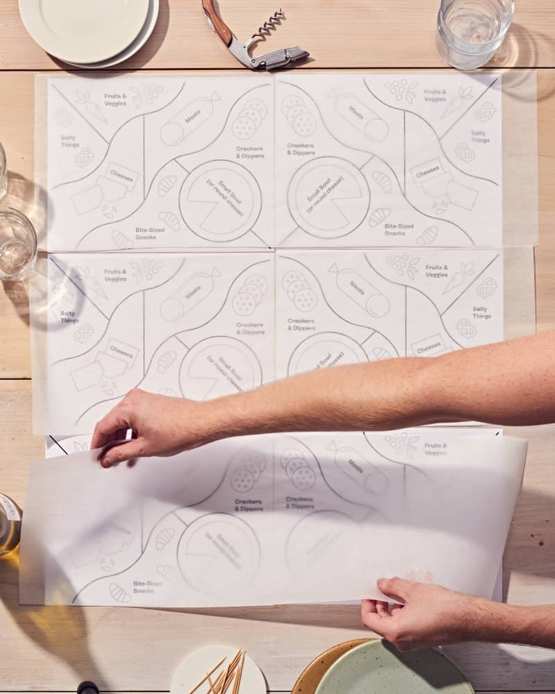 Translucent parchment paper being laid upon a grazing board template that shows you how to place snacks, cheese, meat, fruit etc in order to have a party