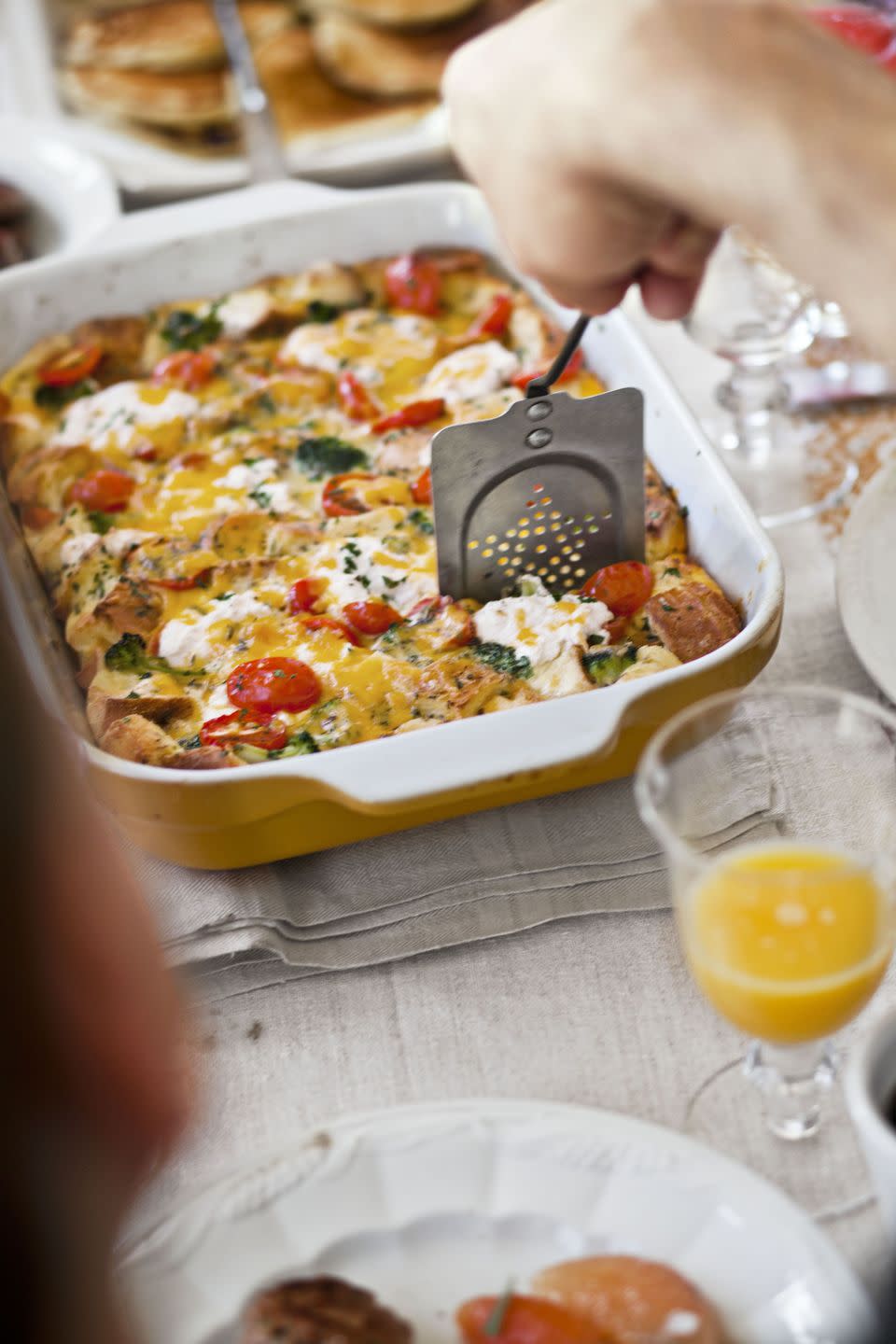 a casserole dish of tomato cheddar strata with broccoli on a table with a hand reaching in to take a slice using an old fashioned spatula