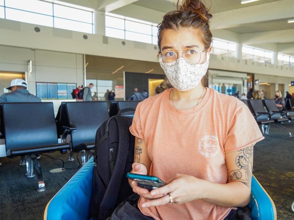 The author sits in a seat at an airport gate with a mask on holding her phone