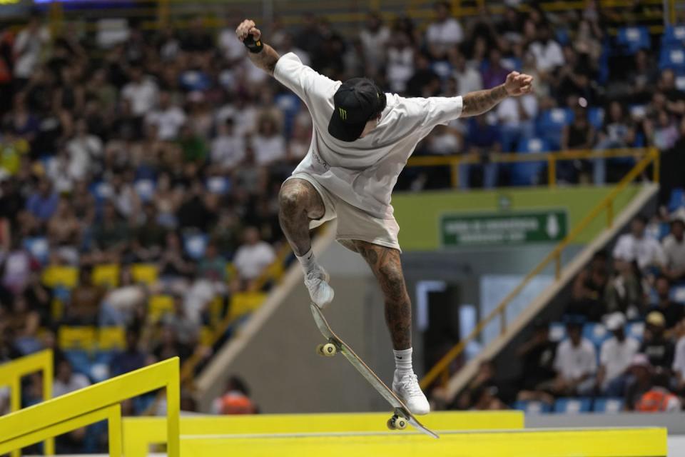 A man in a baseball hat and white shirt and shorts balances mid-air on a skateboard, in front of a seated crowd.