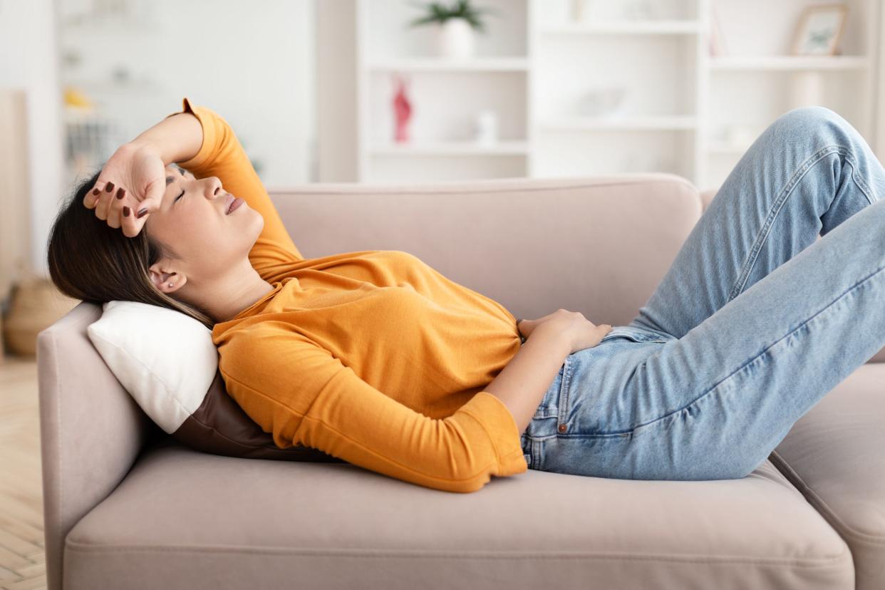 Feeling unwell, menstruation, flu, migraines and illness. Sad despair young asian woman lying on couch suffering from headache and abdominal pain in living room home interior