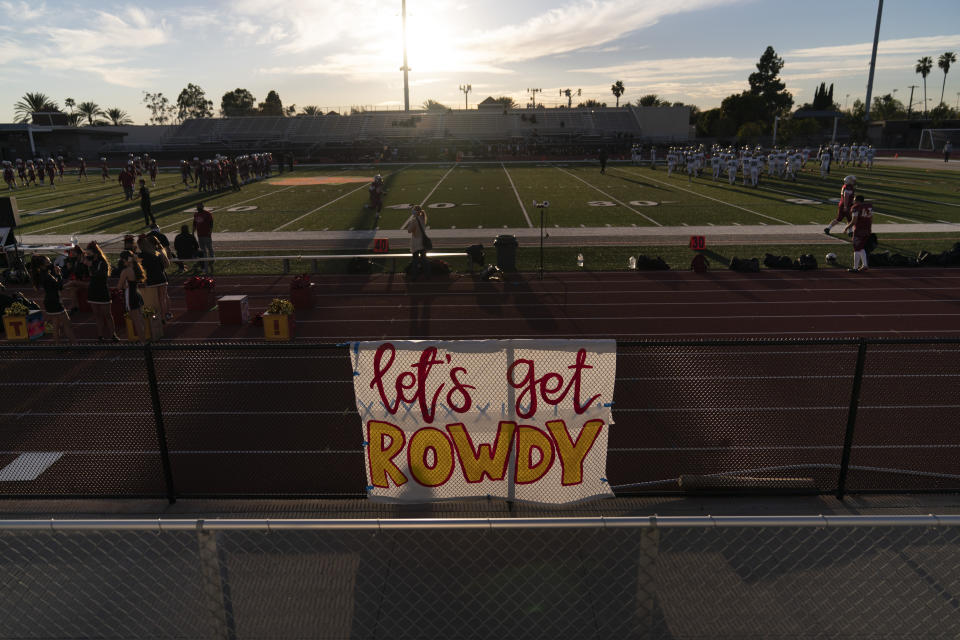 A sign hangs on a fence as players warm up before a high school football game between El Modena and El Dorado in Orange, Calif., Friday, March 19, 2021. The team recently played its first football game in a year as more California counties ease coronavirus restrictions and life in the nation’s most populous state inches back to normalcy. (AP Photo/Jae C. Hong)