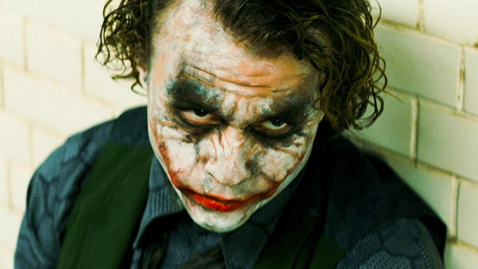 Heath Ledger as the Joker sits against a brick wall.