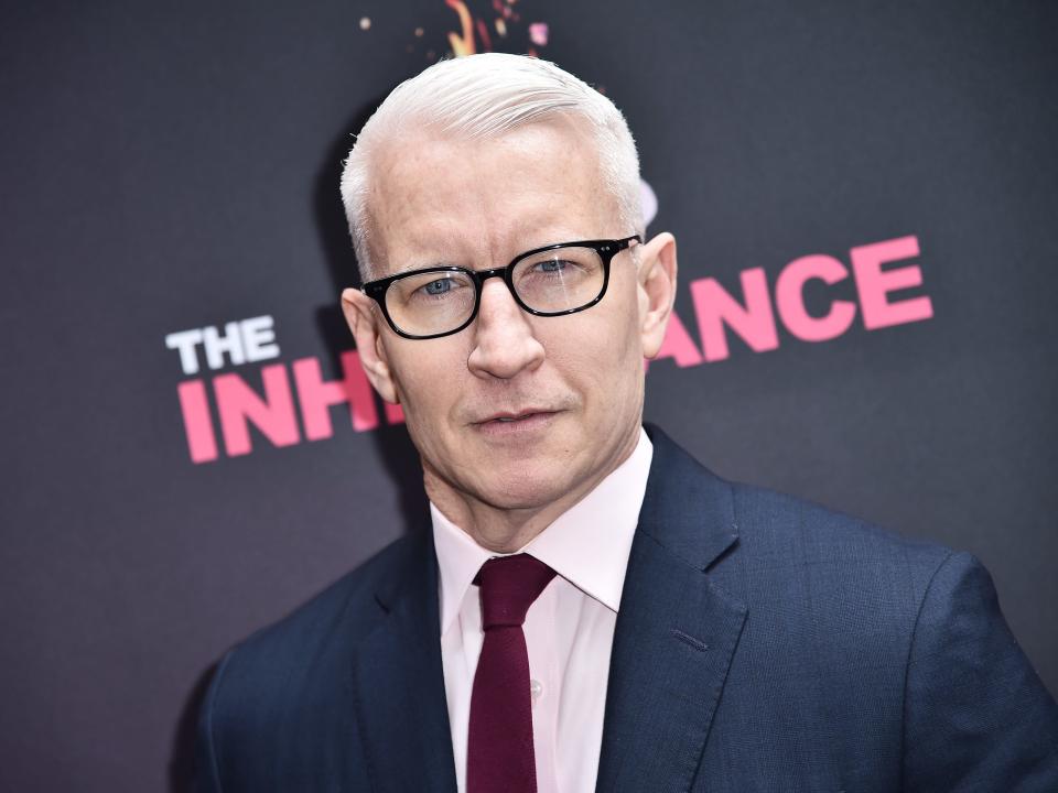 Anderson Cooper at event. He wears glasses, a navy suit, and a pink shirt and tie.