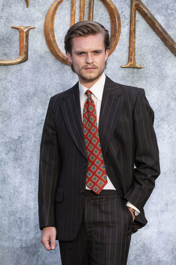 Jack Lowden stands against a backdrop, wearing a dark pinstripe suit with a white shirt and geometric-patterned tie