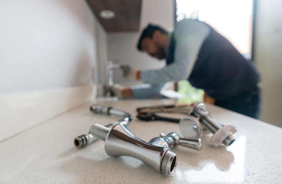 Plumbing parts are seen in the foreground with a man fixing a faucet in the background.