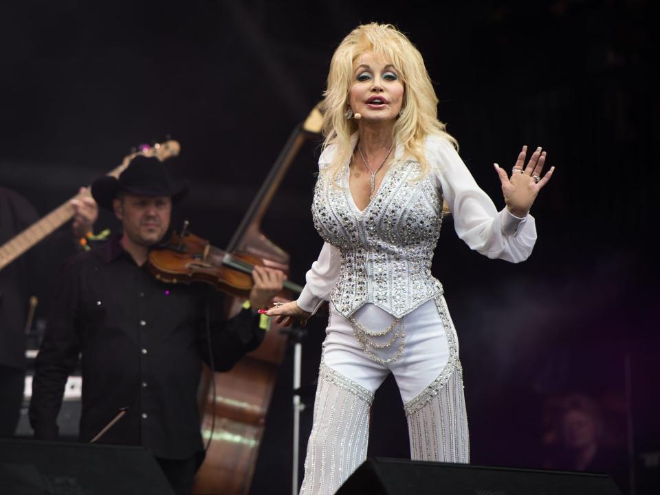 Dolly Parton singing in a white vest and pants covered in silver crystals and mesh.