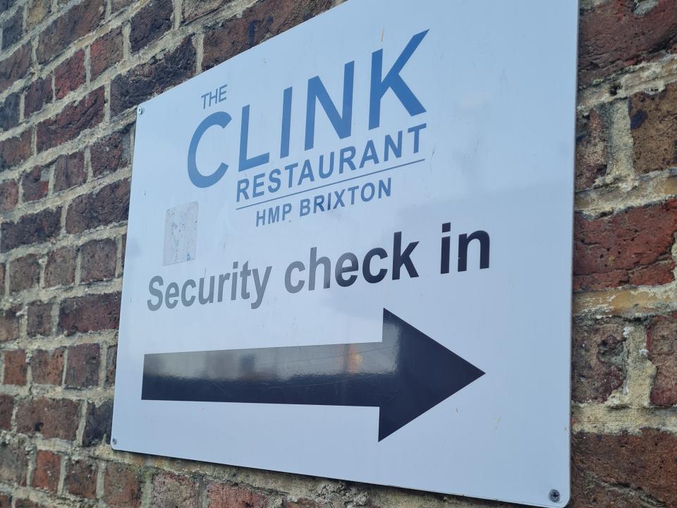 sign posted to the side of a brick wall pointing toward the security check in area for the clink restaurant at brixton prison