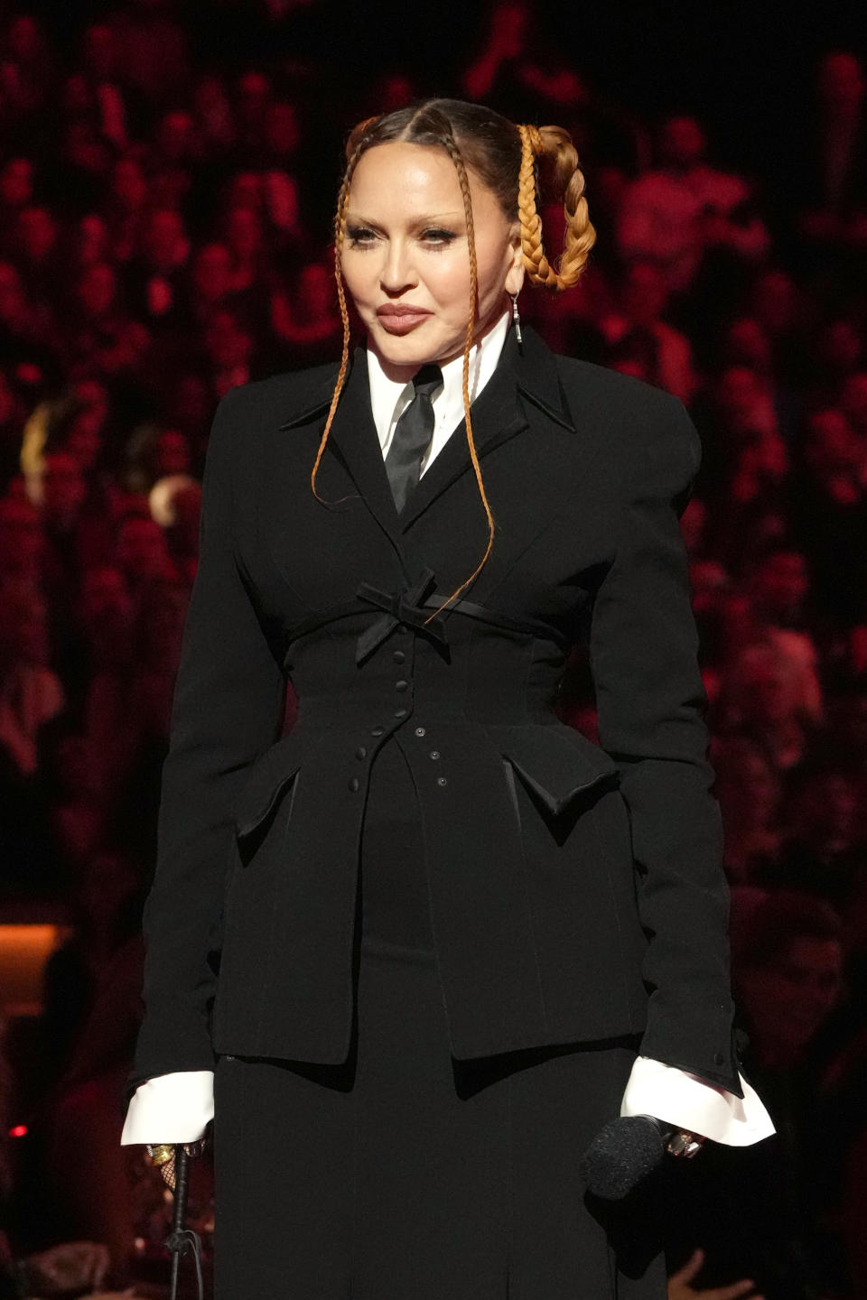 Madonna in a tailored black suit with braided hair at an event, holding a microphone