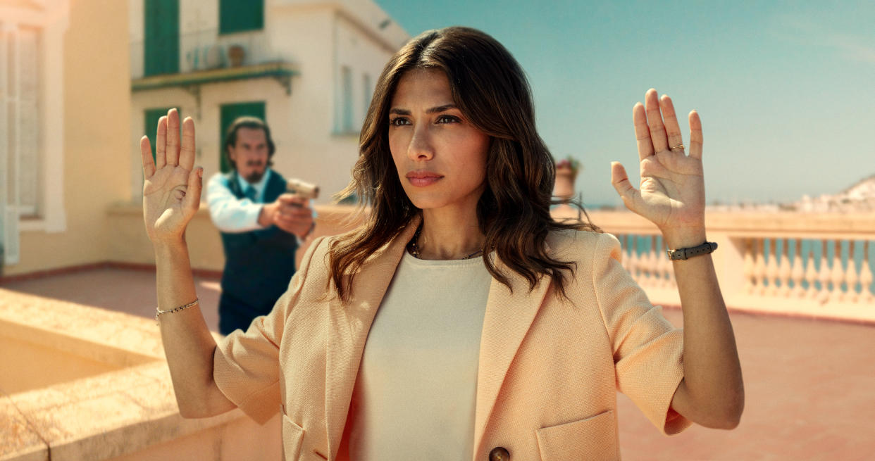  An episodic still from Who Is Erin Carter? showing Erin (Evin Ahmad) standing on a rooftop, holding her hands up while a man behind her is pointing a gun at her 