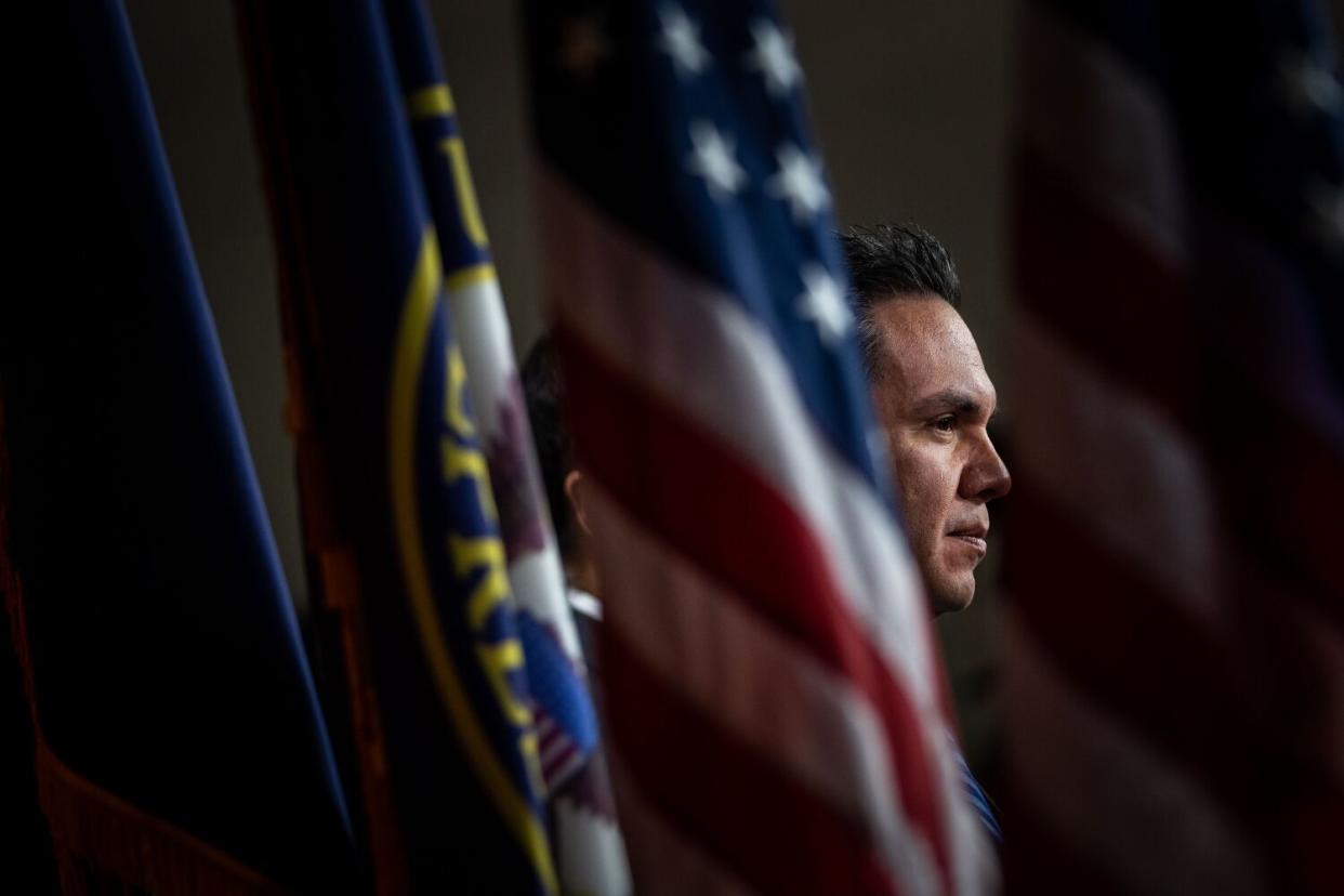 Pete Aguilar is framed head-and-shoulders from a profile, looking toward to right side, standing between flags.