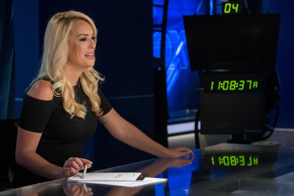 Britt McHenry sits behind a news desk while holding a pen and looking at the teleprompter