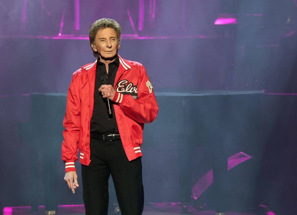 barry manilow stands on a stage in a black shirt and pants with a red jacket