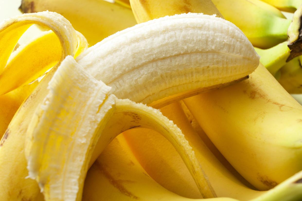 Closeup of an opened banana laid over several unopened bananas, pointing towards the right, with sunlight hitting the top left corner