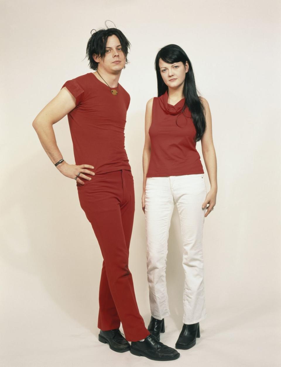 jack white and meg white stand for a studio portrait, he wears a red shirt and matching pants with black shoes, she wears a red shirt and white pants with black shoes