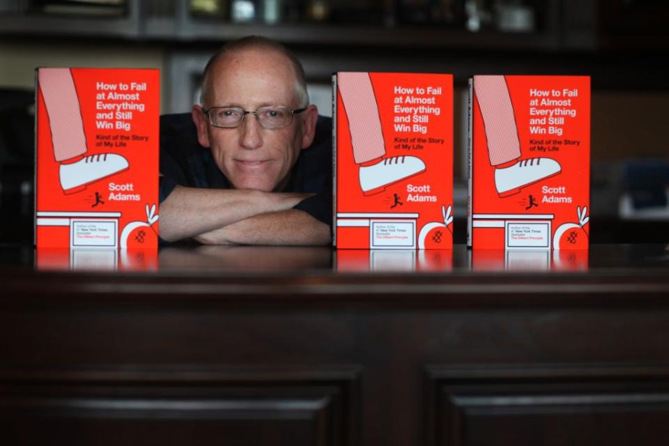 scott adams sitting at desk between copies of his book how to fail at everything and still win big