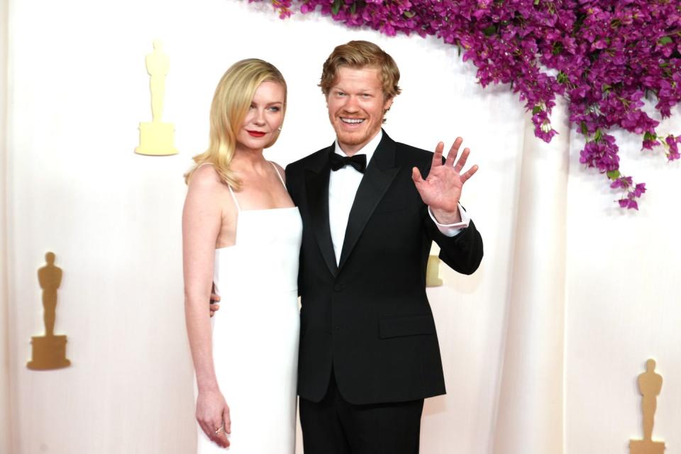 actor jesse plemons waving toward the camera and smiling as he embraces wife kirsten dunst