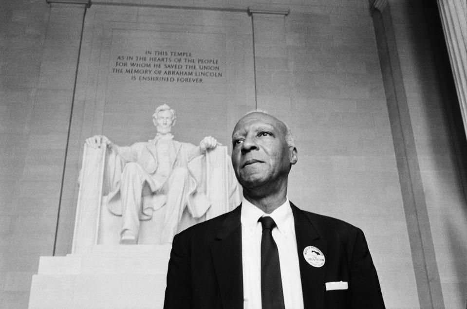 a philip randolph stands in front of a statue of abraham lincoln memorial, randolph wears a suit and tie with a button on his jacket