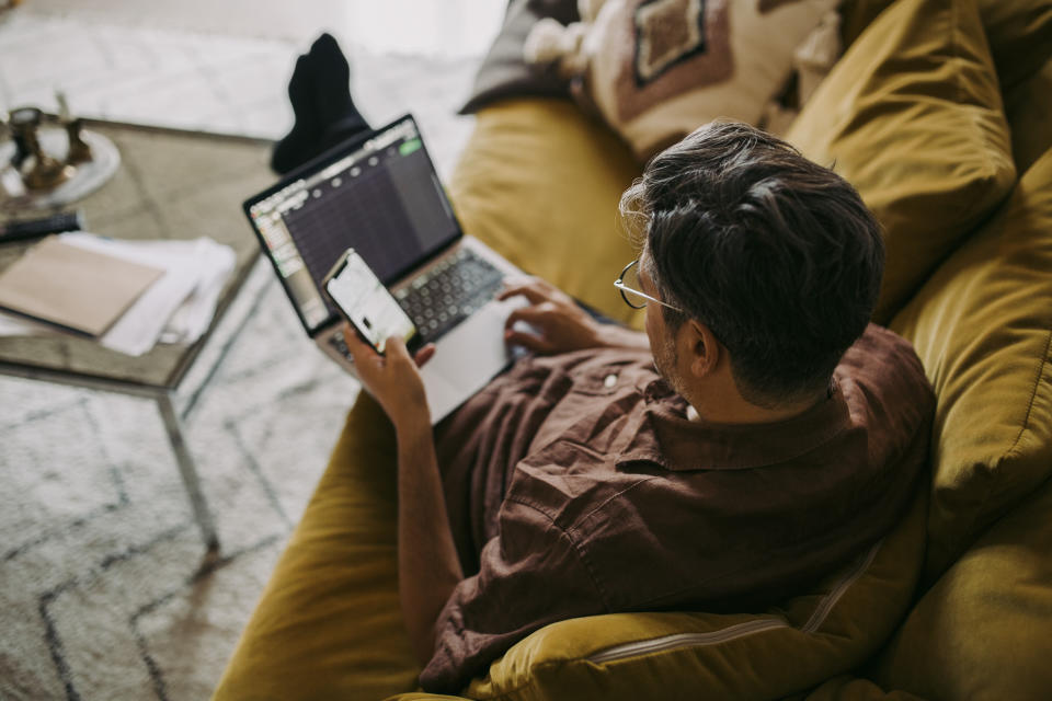 Man lounging on sofa using his laptop and mobile phone simultaneously.