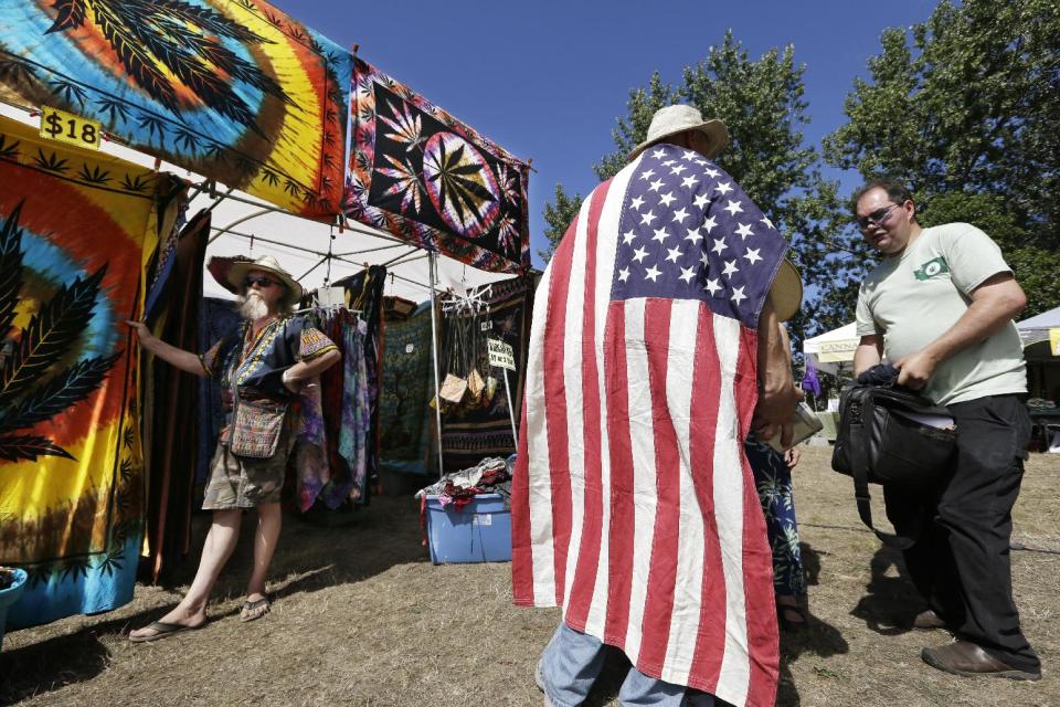 FILE -- In this Aug. 16, 2013 file photo, a man wears an American flag as he walks past a vendor selling tie-dyed products at the first day of Hempfest in Seattle. One of the U.S. Justice Department's top concerns in allowing Washington and Colorado to move forward with plans to legalize and tax marijuana sales is seeing that the states keep criminals out of the industry. But the DOJ itself is refusing to let Washington run national background checks on those applying to run legal pot businesses, The Associated Press has learned, and those who have received the first legal pot licenses have done so without going through a national background check. (AP Photo/Elaine Thompson, File)
